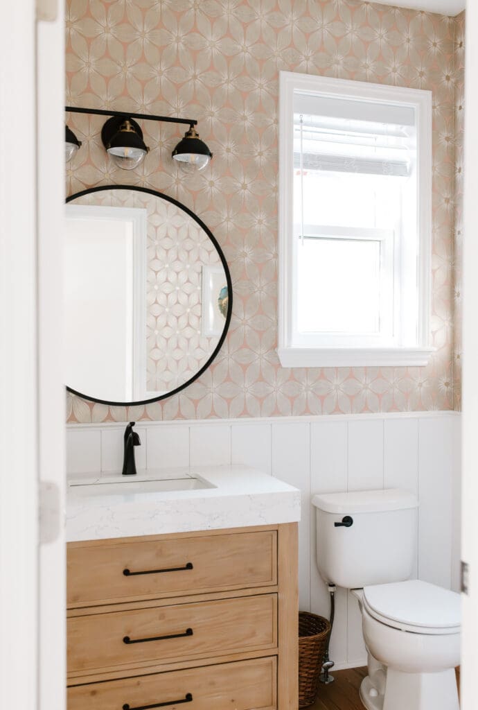 Powder bathroom with wood vanity, white wainscot, metallic wallpaper, and matte black fixtures and round mirror